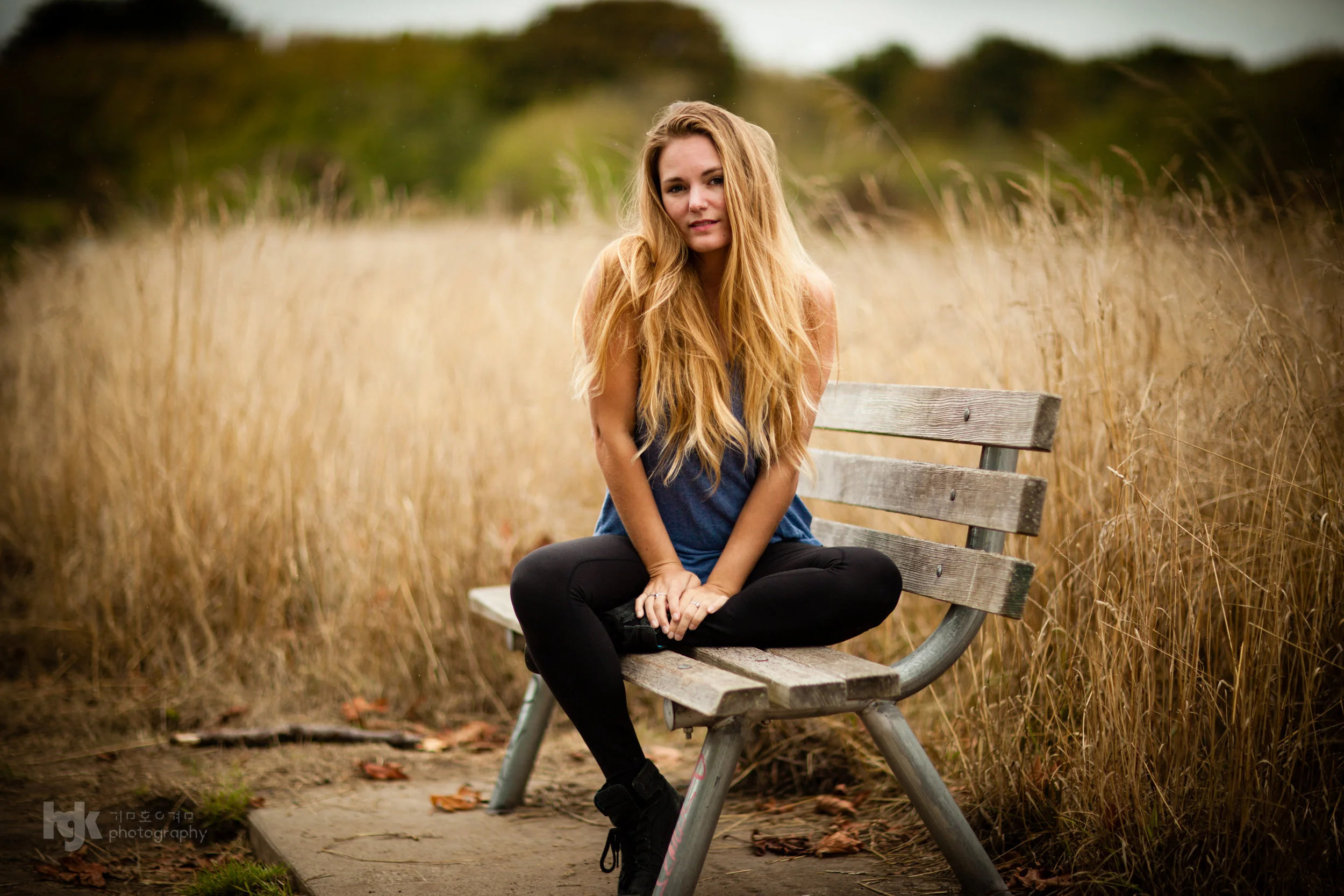 Editorial portrait of a model seated cross-legged on a park bench surrounded by dry grass.