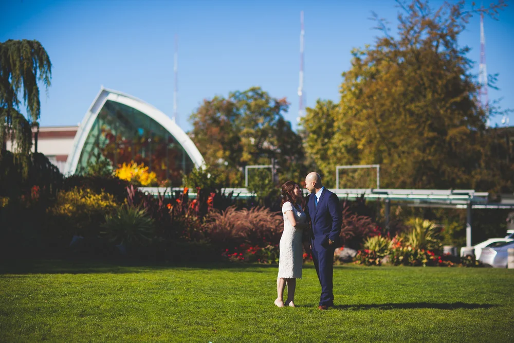 Couple standing together in a landscaped park with an arched building in the background.
