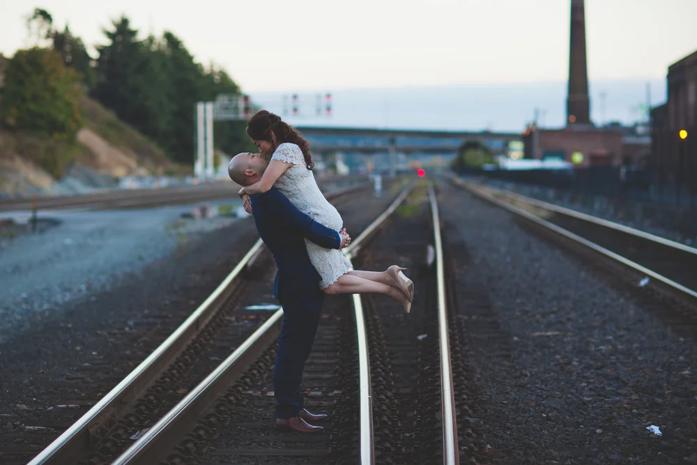 Couple laughing during a playful piggyback pose on railway tracks at dusk.