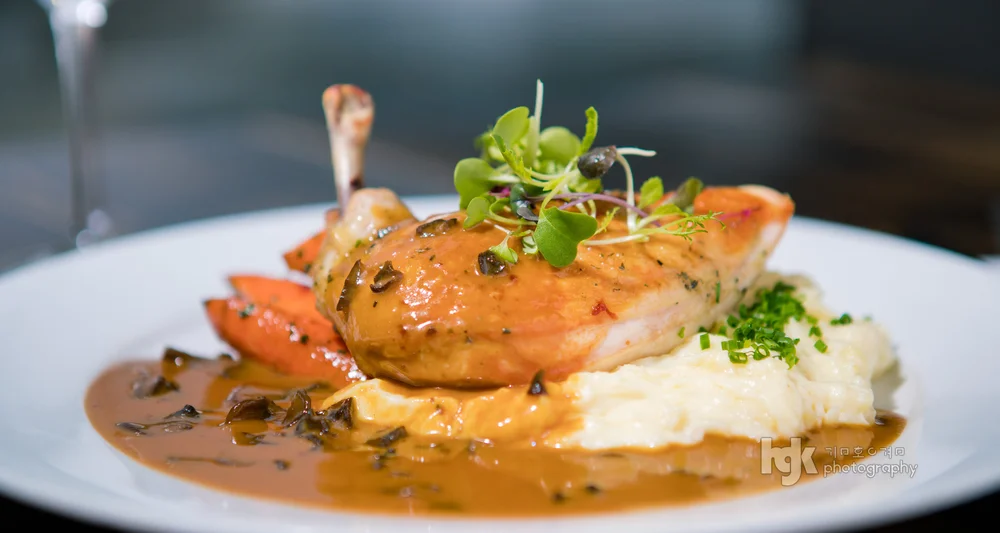 Plated roasted poultry leg in sauce over mashed potatoes with carrots and greens.