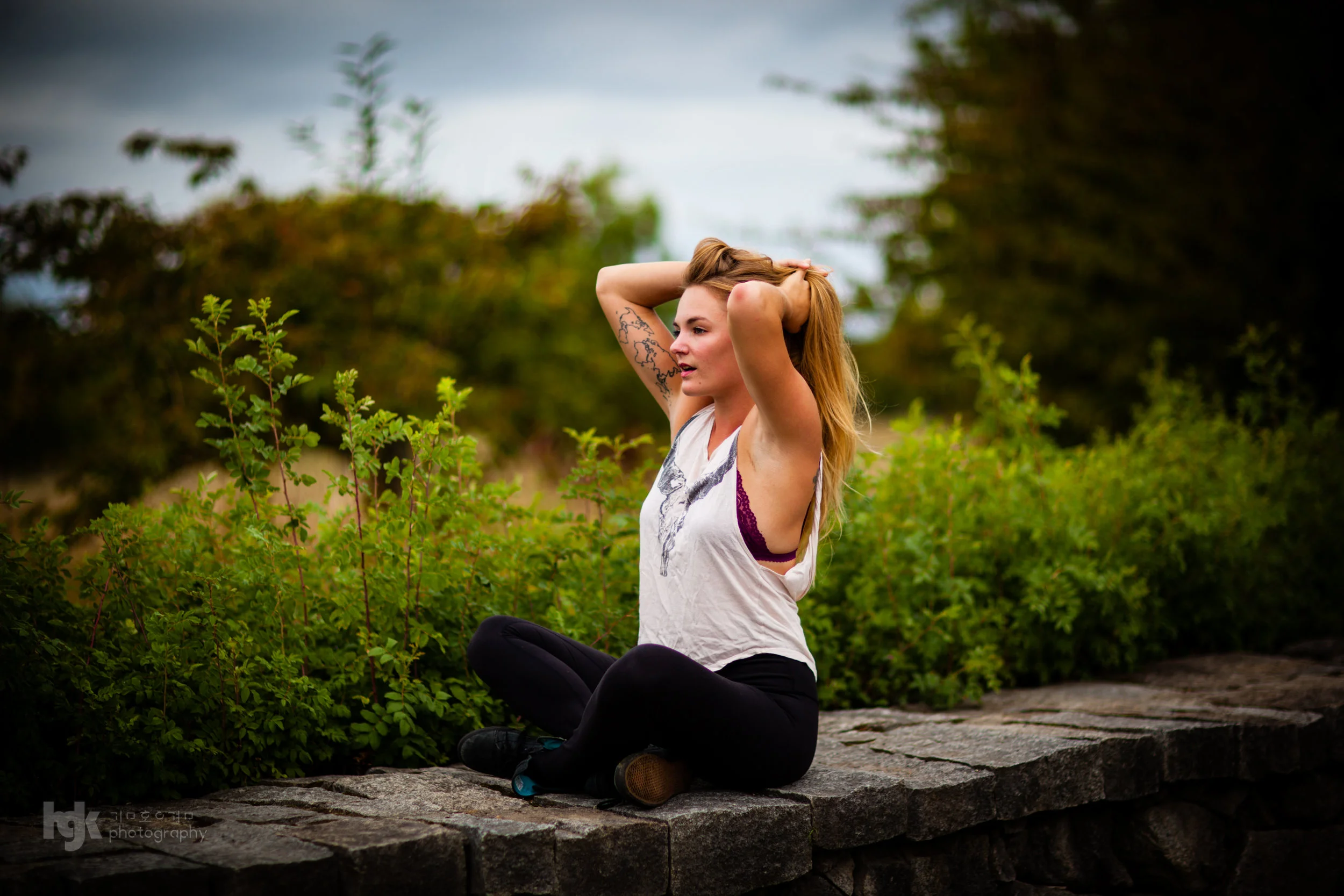 Editorial portrait of a model seated on a low stone wall in a white tank top amid green shrubs.