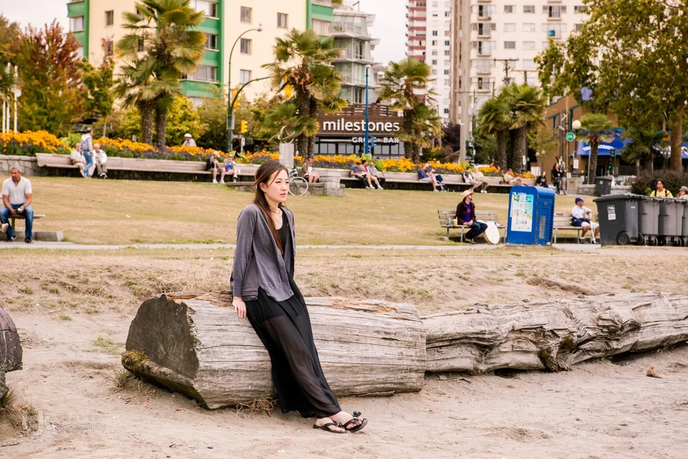 Editorial portrait of a model in dark clothing seated on a large driftwood log in a Vancouver park.