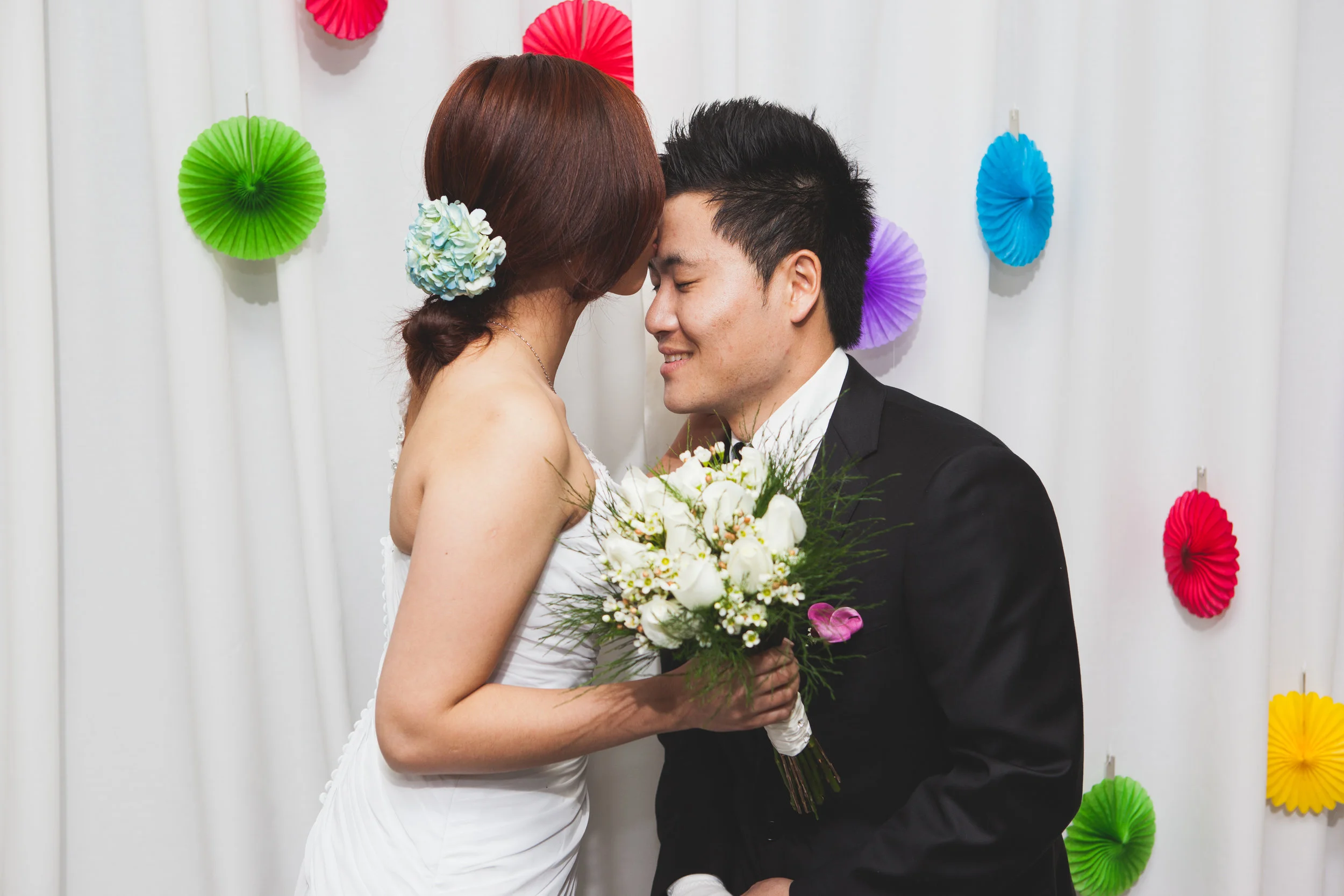 Bride and groom touching foreheads while holding a white bouquet against a colorful backdrop.
