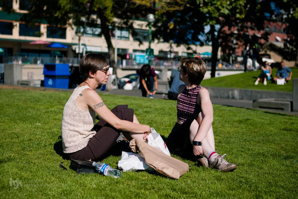 Two people sitting on a city lawn talking beside shopping bags.