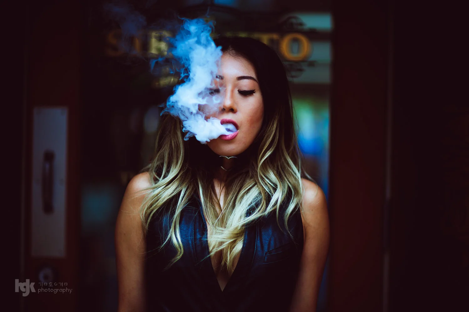 Close editorial portrait of a model exhaling blue-tinged smoke near a storefront doorway.