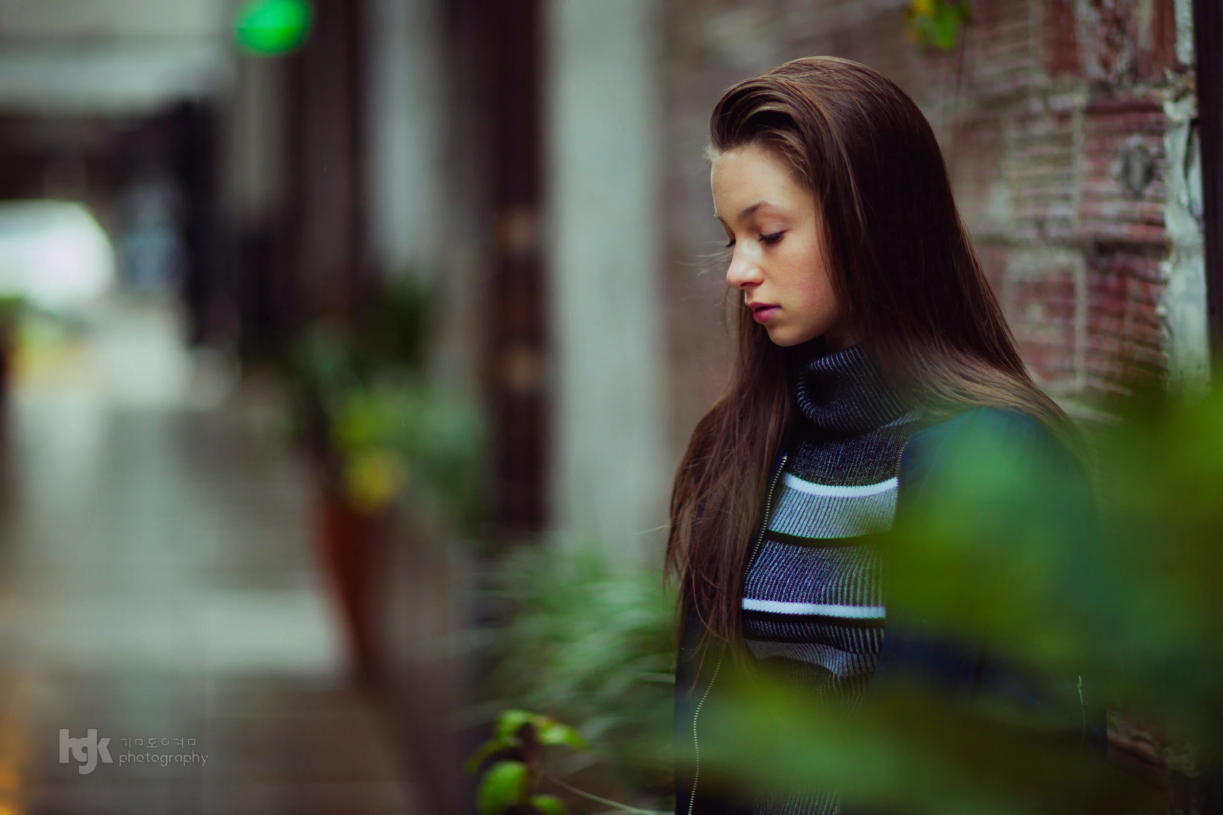 Editorial portrait of a model in layered knitwear, framed through soft foreground greenery.