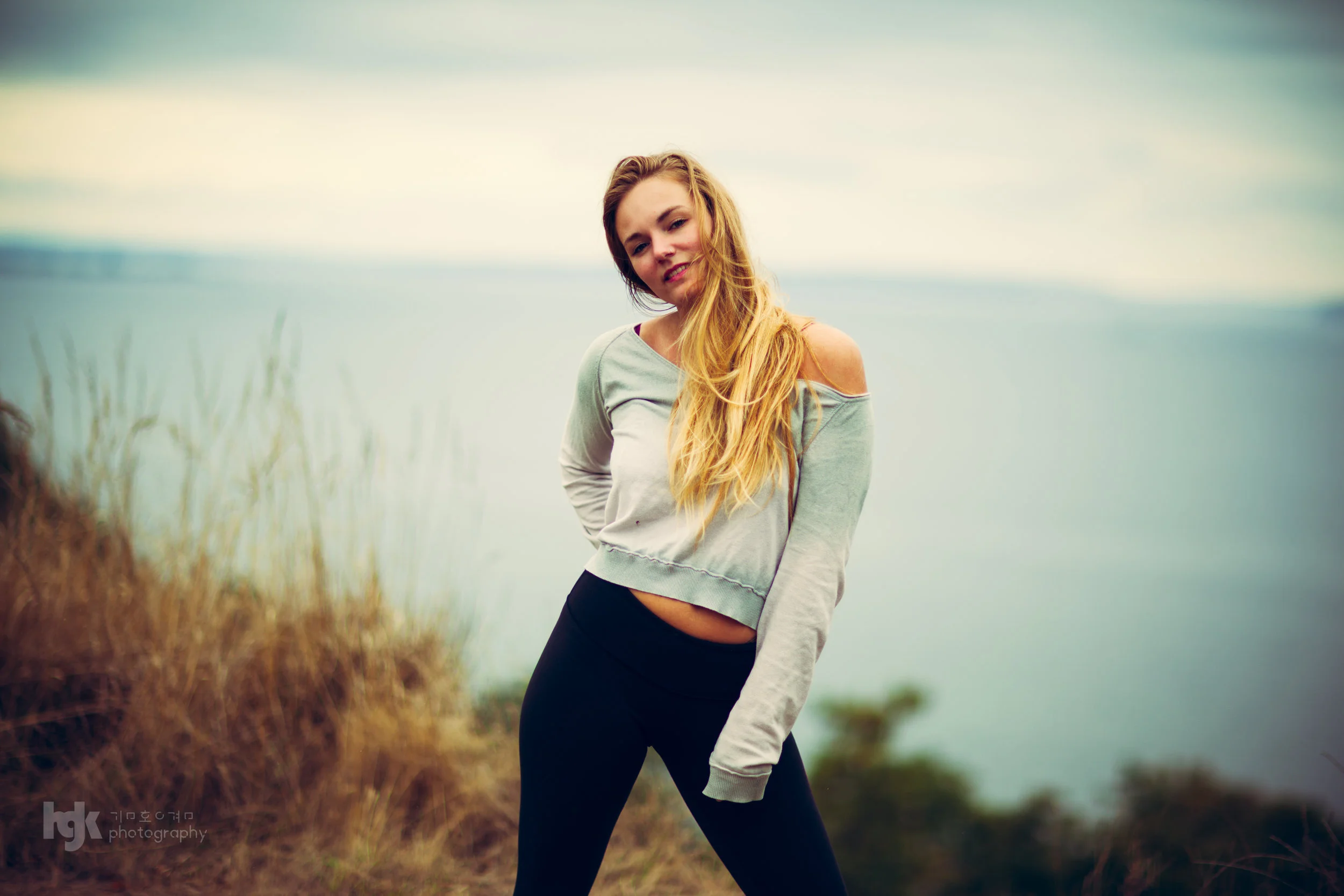 Editorial portrait of a model in a gray off-shoulder top against an overcast waterfront.