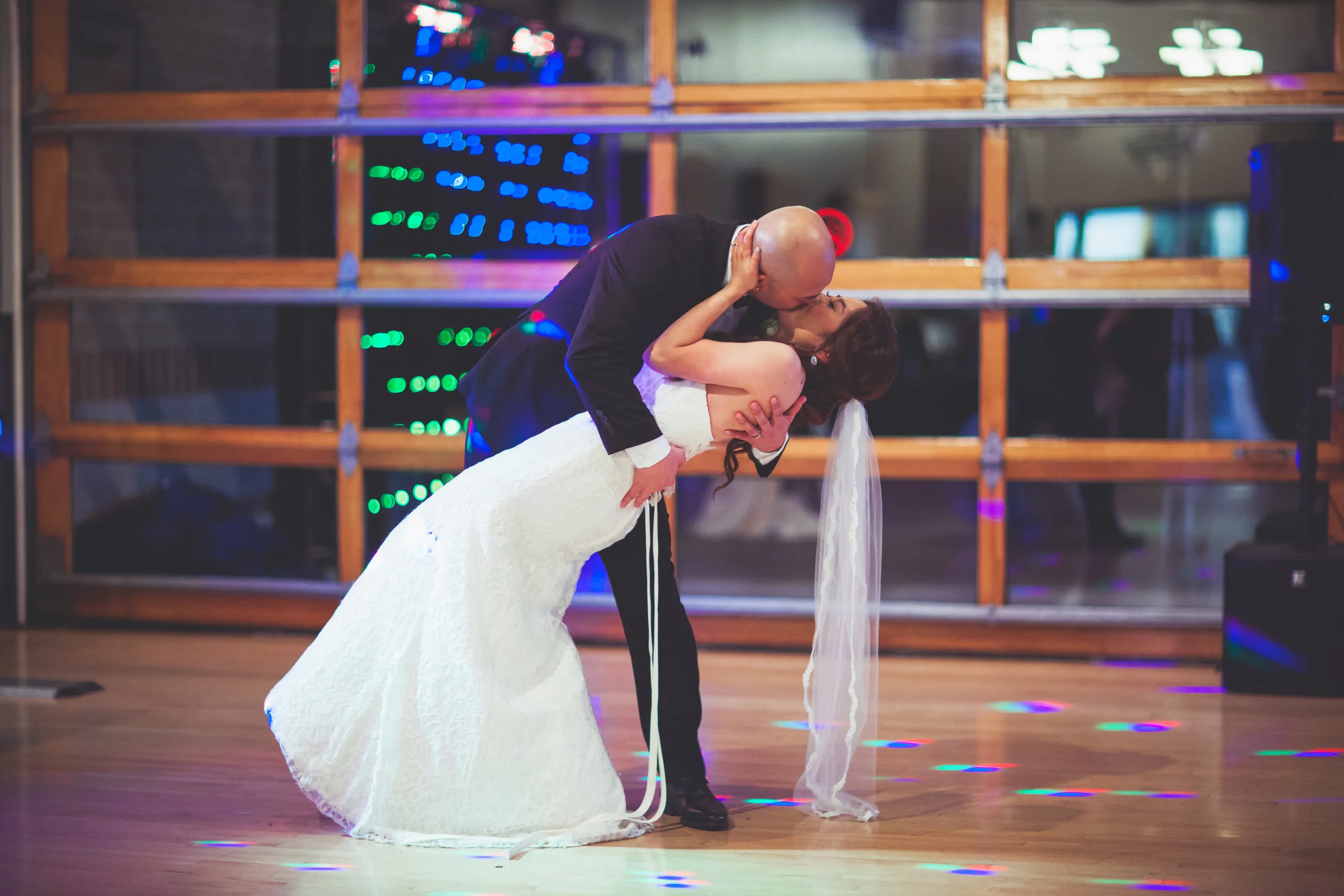 Newlyweds kissing on a dance floor under colorful reception lights.