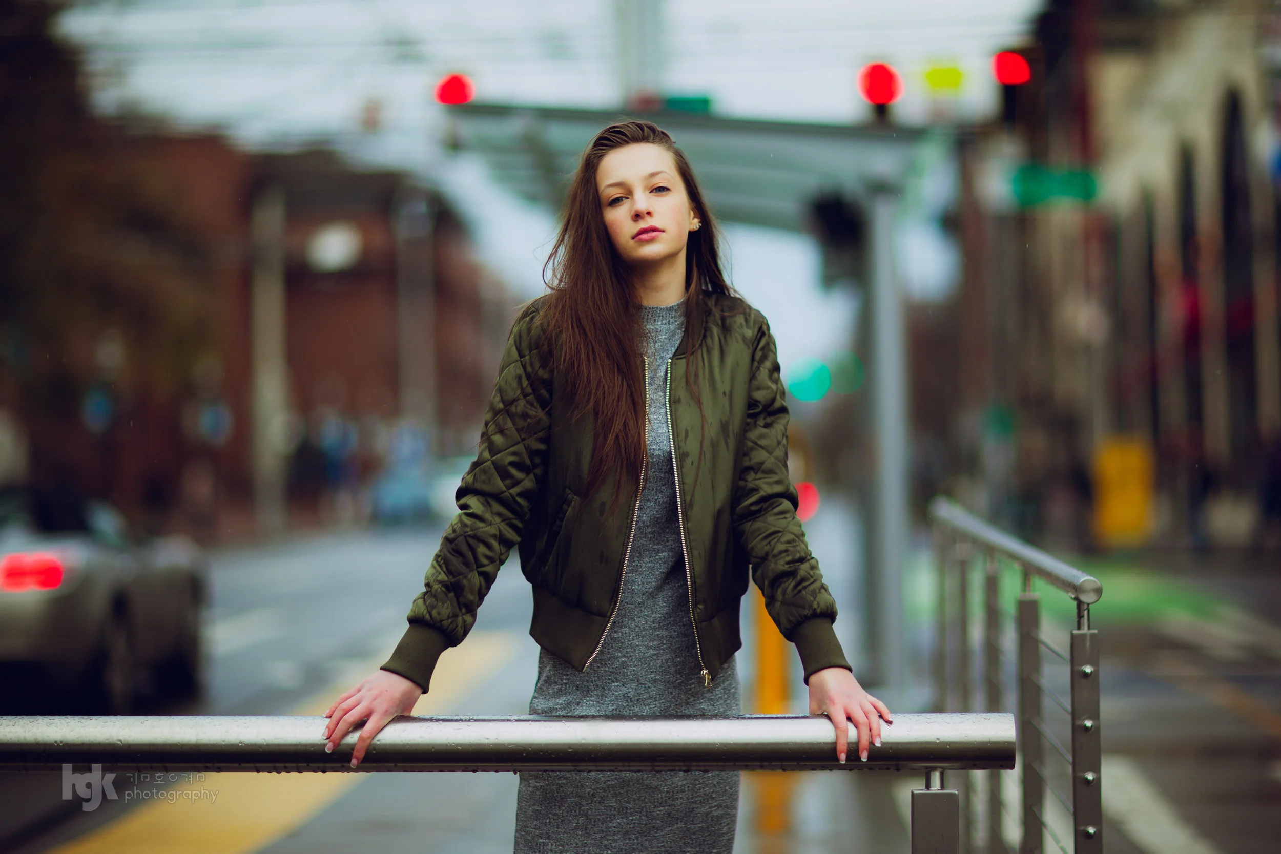 Editorial portrait of a model in an olive bomber jacket and gray dress leaning on a city railing.